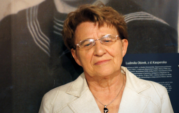 Starsza kobieta w okularach i białej marynarce na tle wielkoformatowego zdjęcia ok 12-letniej dziewczynki w mundurku szkolnym