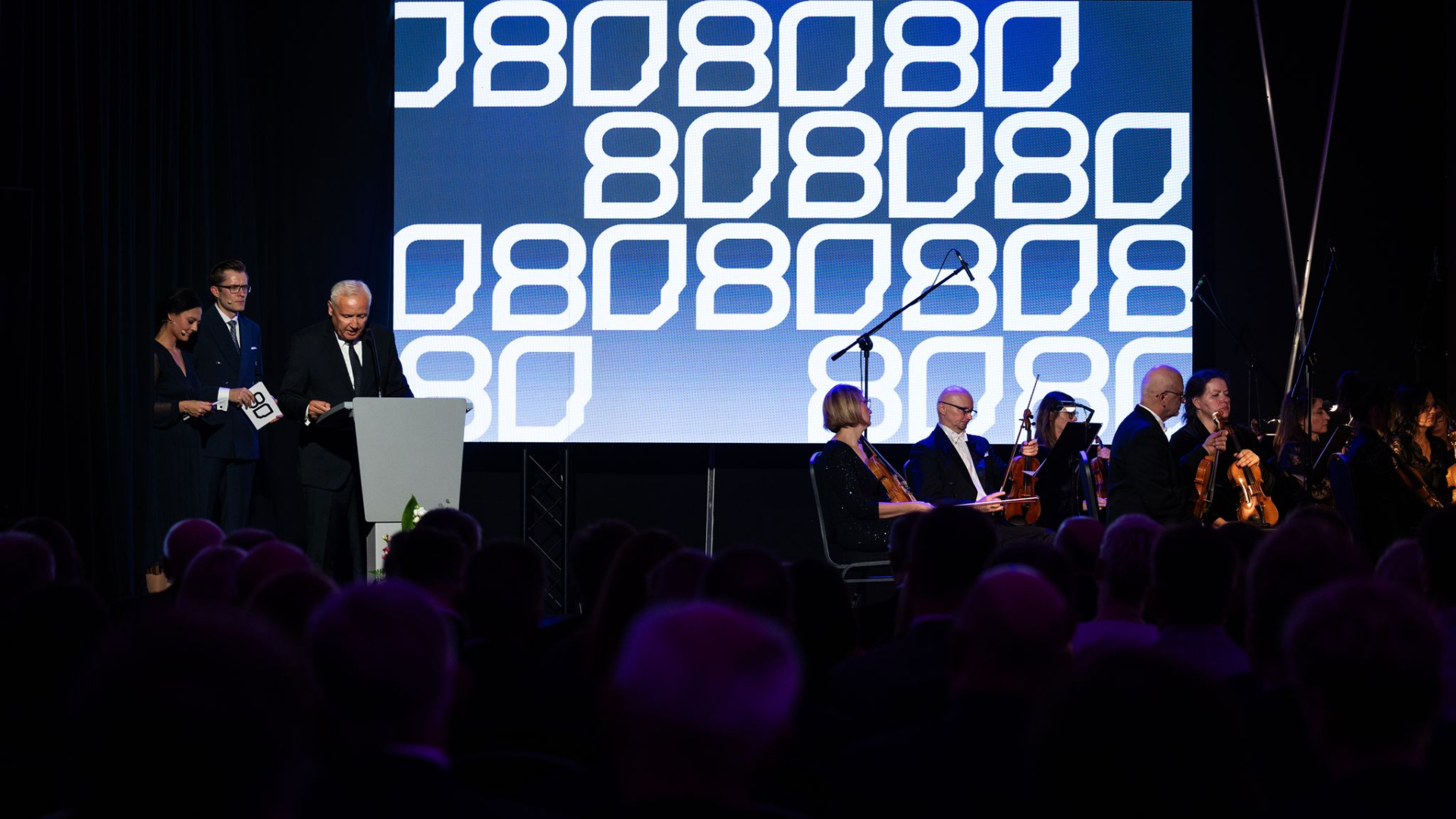 A group of people on stage, the host, the director and a group of musicians, with a screen displaying the number 80 in the background. 