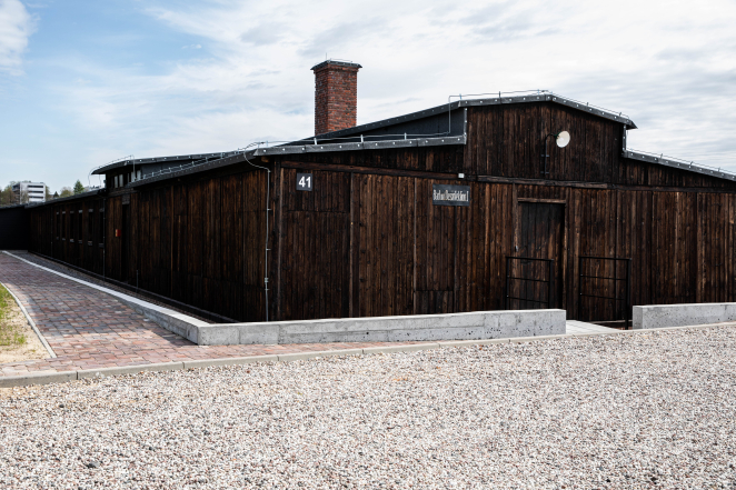 Drewniany barak więźniarski i prostokątnym wysokim kominem, otoczony betonową opaską. 