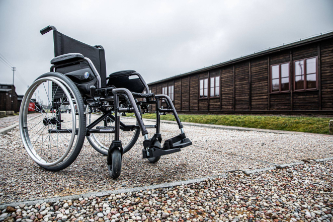 Wózek ortopedyczny ustawiony na drodze wyłożonej drobnym kamieniem, na drugim planie drewniany barak więźniarski z 6 parami okien.