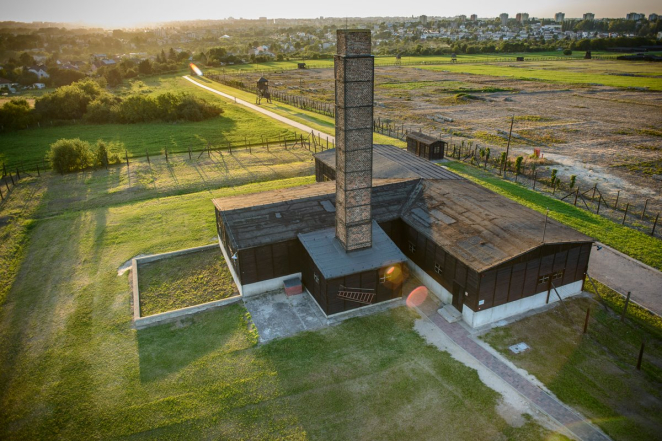 Widok z lotu ptaka na Krematorium, rozłożysty drewniany budynek składający się z czterech elementów oraz wysokiego prostokątnego komina, na drugim planie miejska zabudowa. 