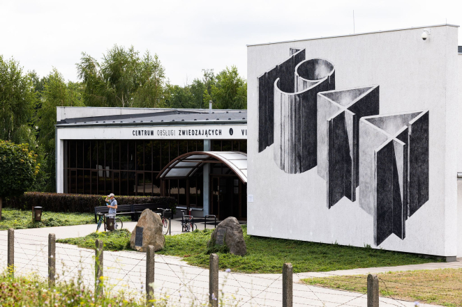 Budynek Centrum Obsługi Zwiedzających, na frontowej ścianie monochromoatyczny mural: 1944, nad wejściem półokrągły daszek, nad nim napis: Centrum Obsługi Zwiedzających, przed obiektem dwa pomniki w kształcie kamieni.