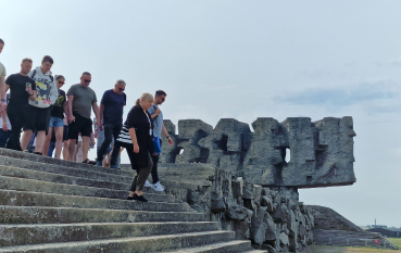 Grupa zwiedzających w letnich ubraniach z muzealną przewodniczką schodzą ze stromych, betonowych schodów, w tle Pomnik-Brama (abstrakcyjna, kamienna forma). 