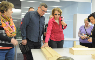 Zwiedzający w przeszklonej sali, kobieta w ciemnych okularach dotyka prawą ręką drewnianego modelu baraku więźniarskiego.