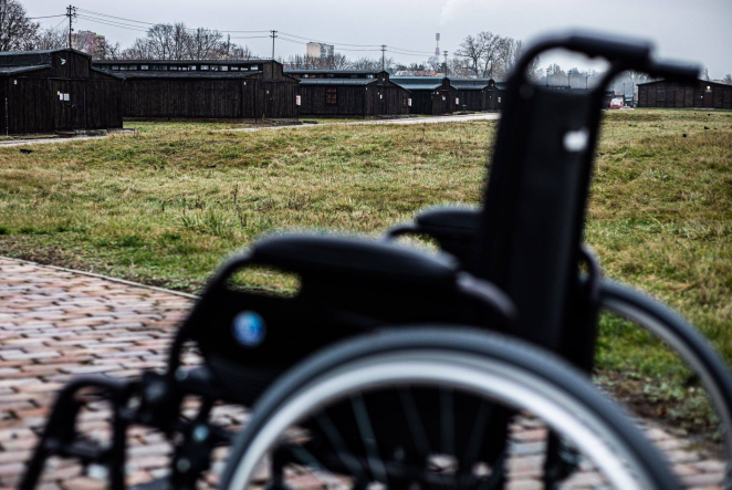 Wózek ortopedyczny na tle rzędu baraków więźniarskich.