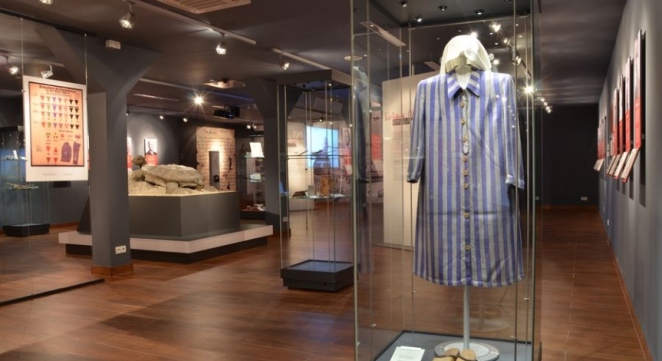 Wnętrze wystawy, szklane gabloty, informacje historyczne na planszach, na pierwszym planie w gablocie sukienka-pasiak oraz biała chusta na głowę, na drugim kamienna rzeźba, żółw