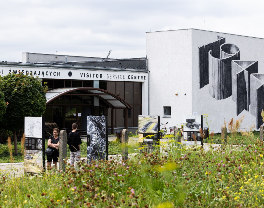 Wejście do Centrum Obsługi Zwiedzających od strony bramy wjazdowej; na pierwszym planie wystawa plenerowa na panelach wystawienniczych oraz oglądający ją ludzie, na drugim wejście do budynku oraz mural: 1944; dookoła zielona trawa i kwiaty polne. 
