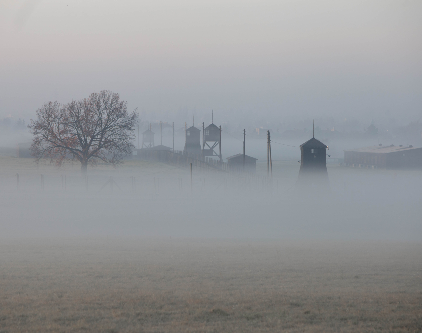 Teren byłego obozu koncentracyjnego na Majdanku spowity przez mgłę, widoczne wieże strażnicze oraz barak więźniarski.