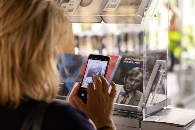 Kobieta o jasnych włosach z telefonem w ręku robi zdjęcie książki Zacheusz Pawlak ustawionej w witrynie księgarni.