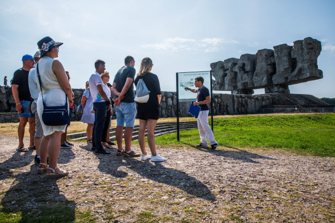 Grupa zwiedzających w letnich ubraniach słucha przewodnika wskazującego na planszę ze ścieżki historycznej, w tle pomnik-Brama.