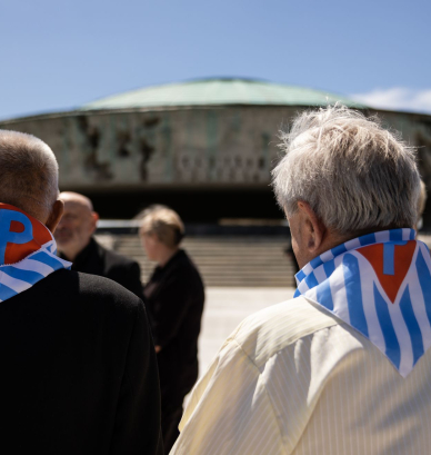 Dwóch starszych mężczyzn z chustami w biało niebieskie pasy z czerwonym trójkątem z literą P stoi na przeciwko Pomnika Mauzoleum 