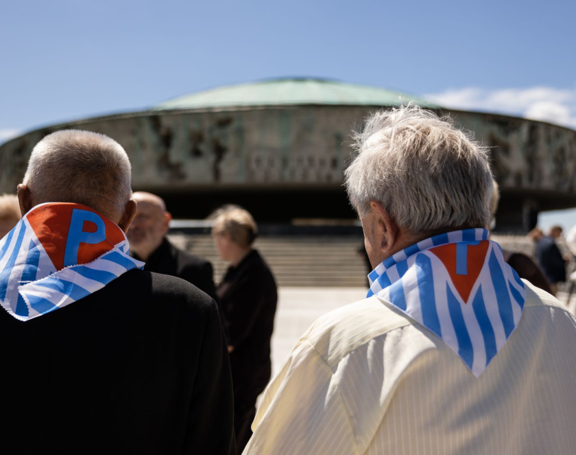 Dwóch starszych mężczyzn z chustami w biało niebieskie pasy z czerwonym trójkątem z literą P stoi na przeciwko Pomnika Mauzoleum
