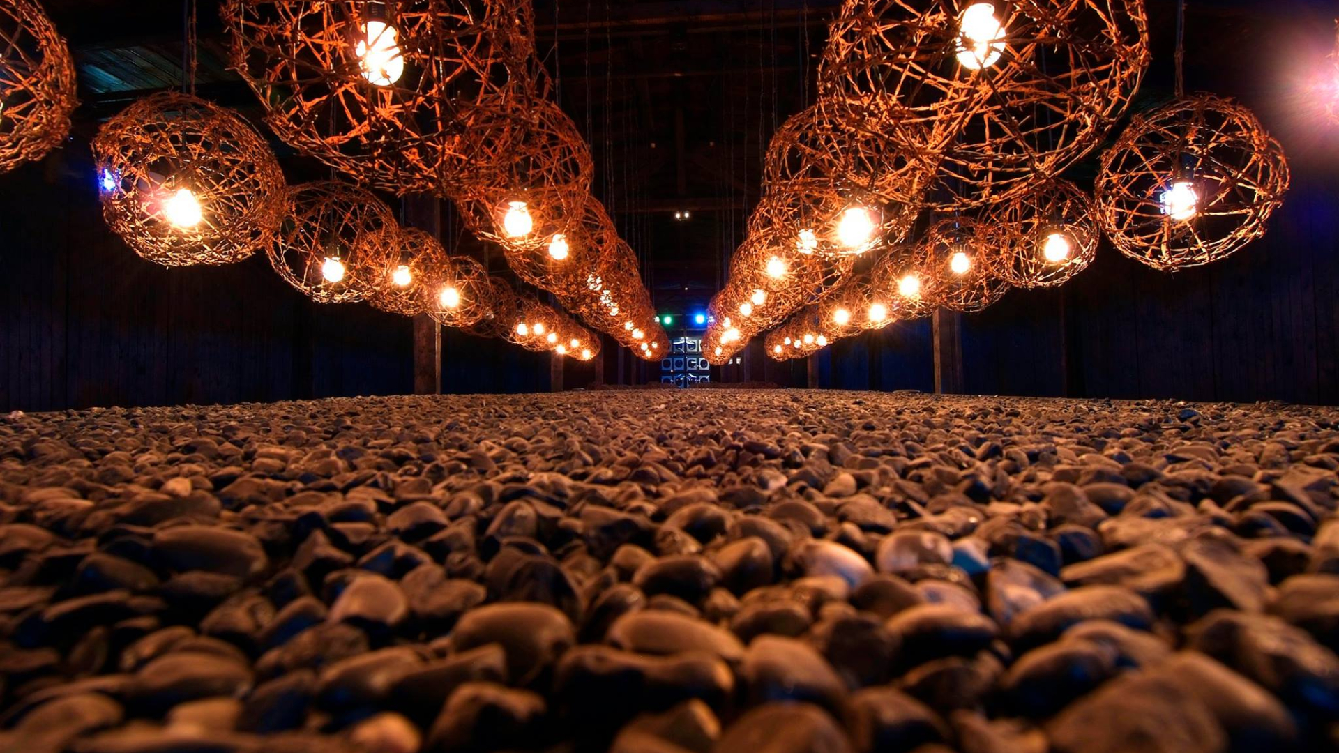 Instalacja artystyczna; cztery rzędy kilkunastu sfer z żarówką w ich centrum zawieszone nad płaszczyzną z drobnych szarych kamieni, dookoła czarna przestrzeń