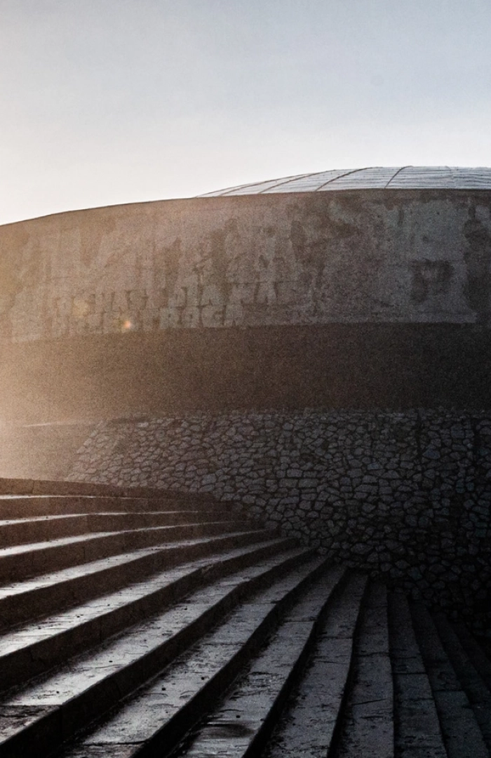 Kopuła Mauzoleum w muzeum na Majdanku widziana od dołu, oświetlony niskim światłem słońca. Na pierwszym planie prowadzą do niego szerokie kamienne schody, a sylwetka budynku odcina się na tle jasnego nieba.