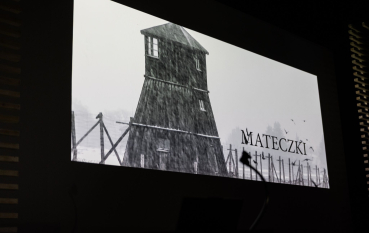 Ciemne pomieszczenie, ekran kinowy, kadr z filmu: wieża strażnicza, padający śnieg, ogrodzenie z drutu kolczastego, kilka wron, czarny napis: Mateczki