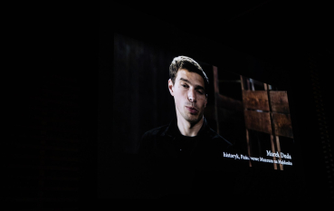 40-letni mężczyzna z czarnymi krótkimi włosami,  na tle prycz więźniarskich, biały napis: Marek Duda, historyk Państwowego Muzeum na Majdanku