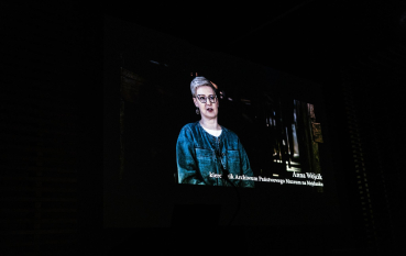 50-letnia kobieta z krótkimi jasnymi włosami w baraku więźniarskim, biały napis: Anna Wójcik, kierownik archiwum Państwowego Muzeum na Majdanku. 