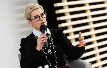 50-letnia kobieta z krótkimi jasnymi włosami, obrana w czarny sweter, sznur pereł oraz białą koszulę z wystającym kołnierzykiem