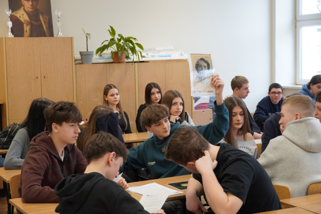 Młodzież szkolna w sali lekcyjnej; w centralnej części nastoletni chłopak podnosi do góry czarno-białe zdjęcie kobiety. 