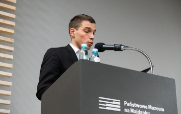 40-letni mężczyzna stojący przy mównicy z mikrofonem, na mównicy dwie butelki wody mineralnej.