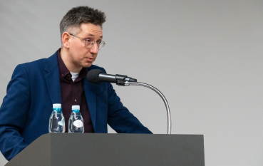 40-letni mężczyzna w okularach przy mównicy z mikrofonem, obok dwie butelki wody mineralnej.