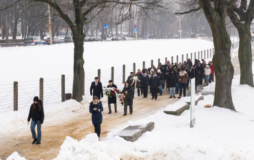 Grupa ludzi niosąca wieńce, po prawej ogrodzenie z drutu kolczastego, po lewej betonowe ławki i drzewa.