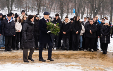 Grupa ludzi, dwie osoby (kobieta i mężczyzna) ubrani na czarno składają kwiaty.