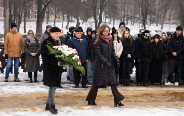 Grupa ludzi, dwie osoby (kobieta i mężczyzna) składają kwiaty.