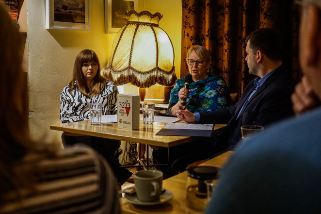 Dwie kobiety i jeden mężczyzna siedzący przy stoliku z ustawioną pionowo książką, za nimi lampa nocna z dekoracyjnym abażurem. 