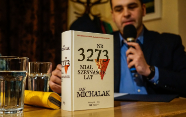 Na pierwszym planie książka, biała okładka czerwony trójkąt, napis: NR 3273 miał szesnaście lat, Jan Michalak, na drugim planie mężczyzna w niebieskiej koszuli mówiący do mikrofonu.