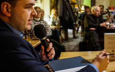 Mężczyzna przemawiający do mikrofonu, na drugim planie grupa osób siedzących przy stolikach.