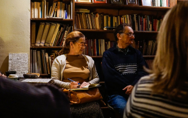 Kobieta i mężczyzna, oboje w okularach, siedzący przy regale z książkami, kobieta robi notatki w otwartym zeszycie. 