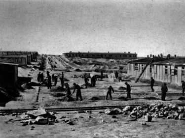 Grupa kilkunastu mężczyzn z narzędziami przy pracy. Po bokach baraki