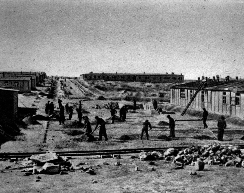 A group of several men with tools at work. Barracks on the sides.