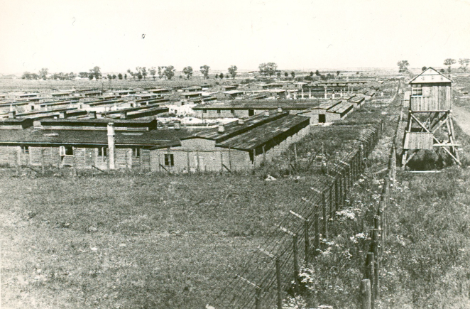 Wooden single-story barracks with windows. On the right, a double wire fence, and behind it a wooden guard tower.