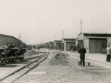 Udeptana ziemia na niej położone tory, kilka wagoników, po lewej i prawej stronie drewniane baraki, po prawej pracujący mężczyźni 