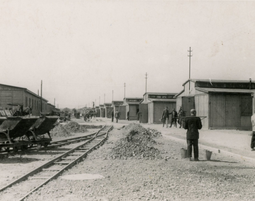 Udeptana ziemia na niej położone tory, kilka wagoników, po lewej i prawej stronie drewniane baraki, po prawej pracujący mężczyźni