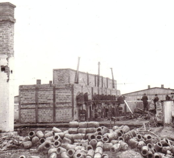 Po lewej stronie ceglany komin. Na środku ceglane piece z metalowymi drzwiczkami. Przed nimi stos cylindrycznych urn.