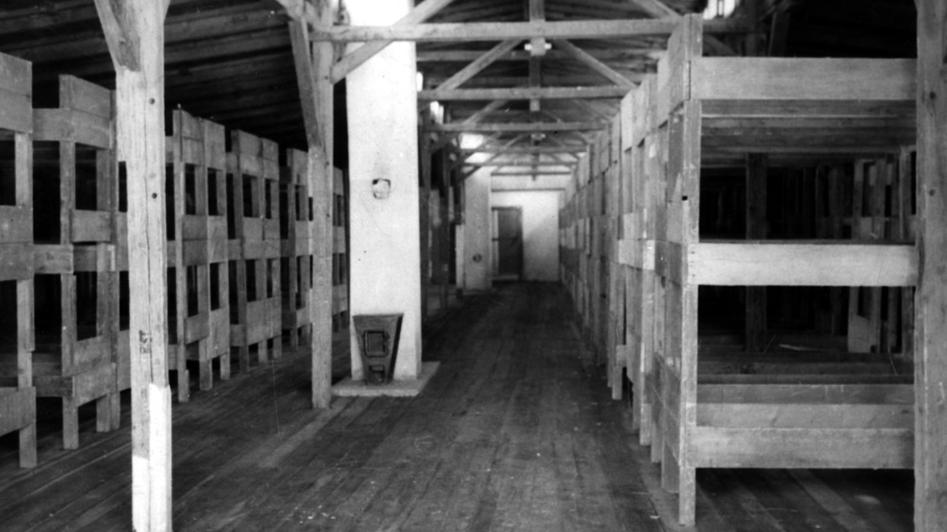 Czarno-białe zdjęcie, wnętrze baraku więźniarskiego, po lewej i prawej strony rzędy piętrowych łóżek. 