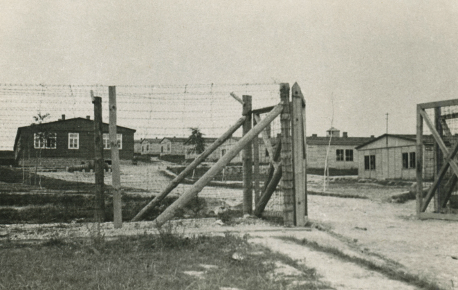 Ogrodzenie z drutu kolczastego z otwartą bramą. Za nim rzędy parterowych drewnianych budynków