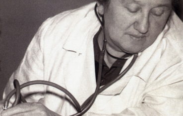 Kobieta w starszym wieku, ubrana w biały kitel i uczesana na gładko. Siedzi przy stole i mierzy ciśnienie innej osobie, której widać tylko rękę.
