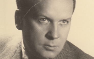 Mężczyzna w starszym wieku, gładko zaczesany do tyłu. Ma na sobie marynarkę, koszulę z kołnierzykiem i krawat.