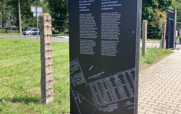 Panel wystawy, napis: Przestrzenie Pamięci, na drugim planie ogrodzenie z drutów kolczastych 