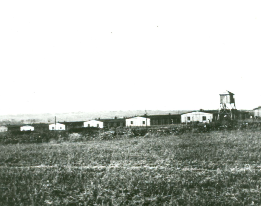 Zdjęcie prezentuje rząd sześciu baraków z oknami. Między nimi stoi wieża wartownicza