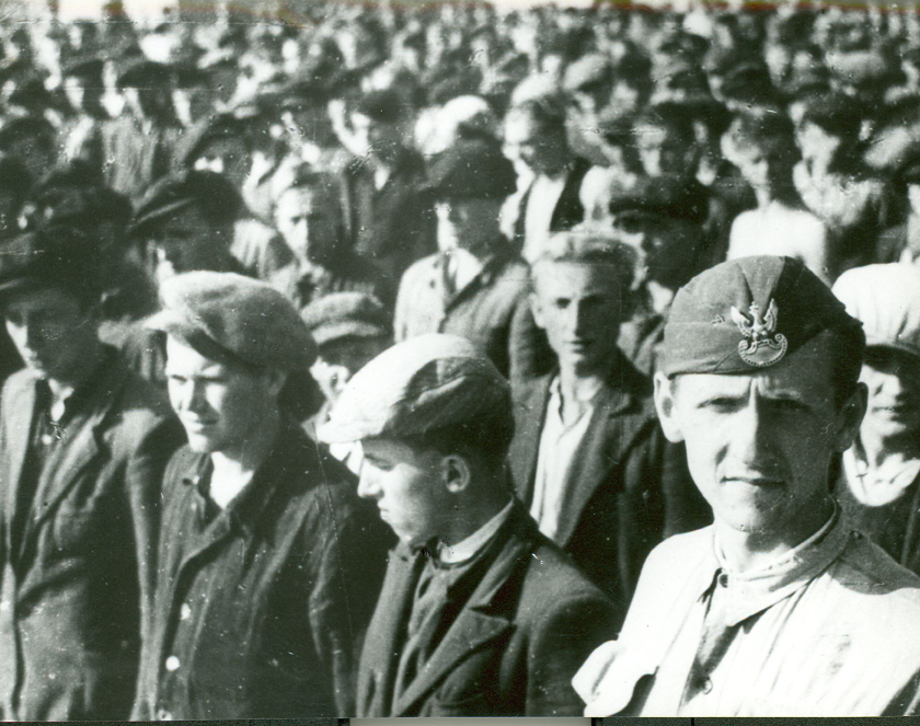 Grupa kilkudziesięciu mężczyzn w cywilnych ubraniach i czapkach ustawionych w rzędach