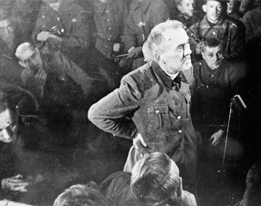 Starszy mężczyzna w niemieckim mundurze stoi wśród innych mężczyzn. Ma założone ręce na biodra i mówi