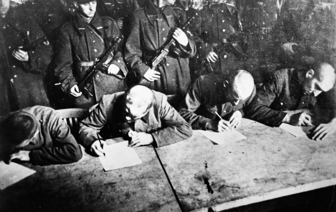 Przy stole siedzi czterech mężczyzn. Trzymają w dłoniach długopisy i pochylają się nad kartkami papieru. Za nimi rząd umundurowanych mężczyzn z bronią w rękach