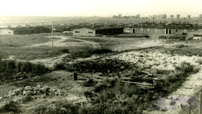 Na pierwszym planie zdjęcia nieużytki i wydeptana ziemia. Dalej kilka parterowych budynków z drewna