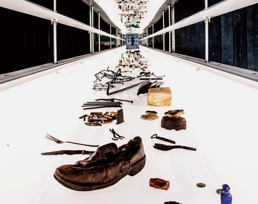 Podniesione wieko szklanej gabloty ukazujące przedmioty, na pierwszym planie męski but