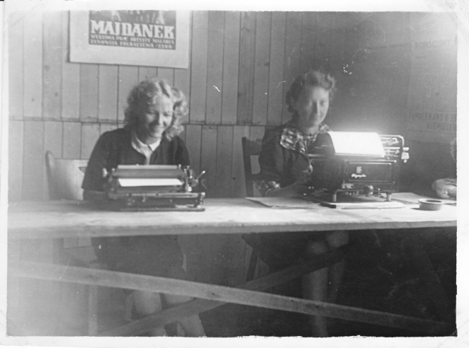 Na zdjęciu dwie uśmiechnięte kobiety. Siedzą przy stole, przed maszynami do pisania. Za nimi plakat z napisem Majdanek
